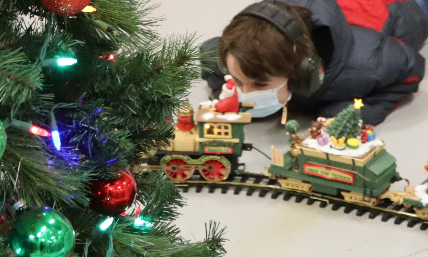 Boy looks at toy train set under a Christmas tree.